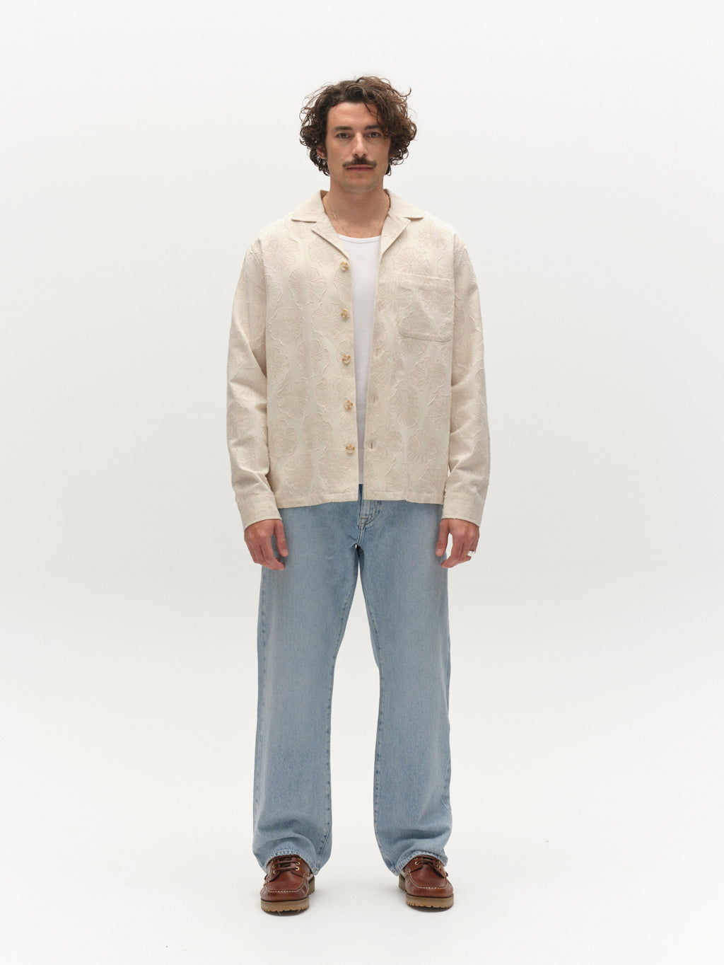 A man with curly hair and a mustache stands against a plain white background, wearing the Gabba GABaustin Reso Raw LS Overshirt in Lt. Sand over a white T-shirt, paired with light blue jeans and brown shoes.