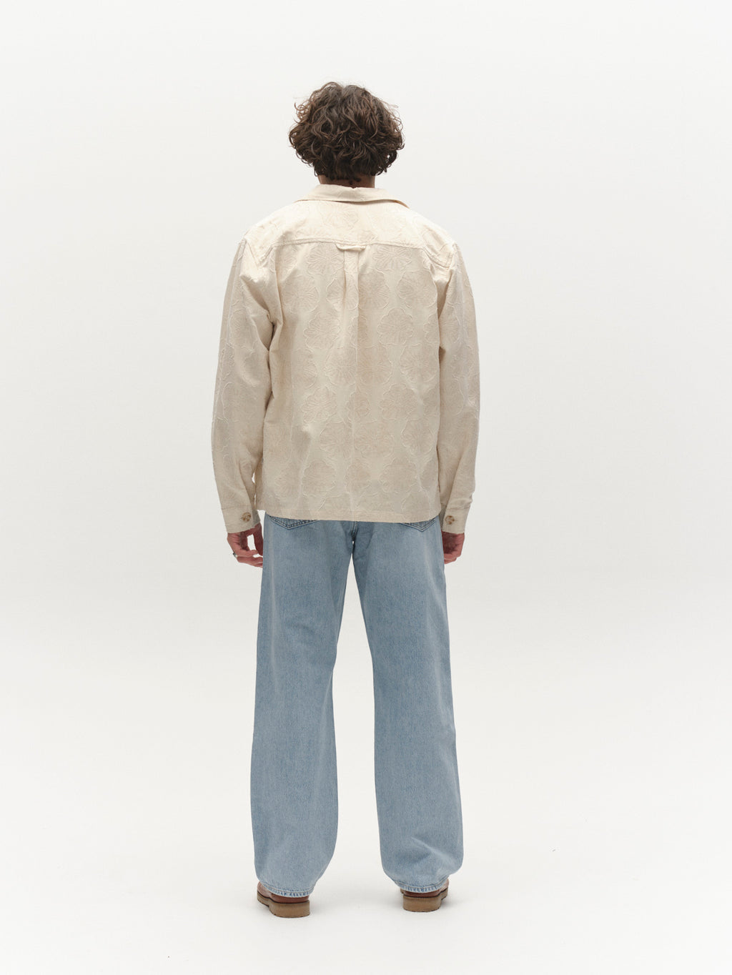 A person with curly hair stands facing away, wearing Gabba's GABaustin Reso Raw LS Overshirt in Lt. Sand and loose light blue jeans, set against a plain white background.
