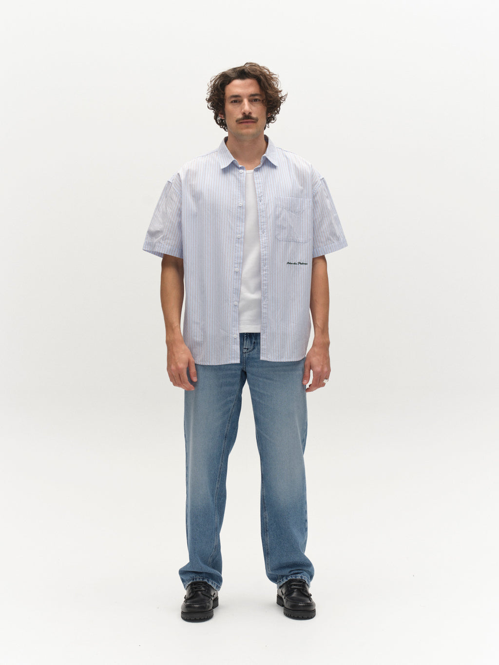 A man with a mustache stands facing forward against a plain white background, wearing a light blue short-sleeve button-up shirt over a white t-shirt, Gabba GABmax Elvis Jeans in Mid Blue Denim, and black shoes.