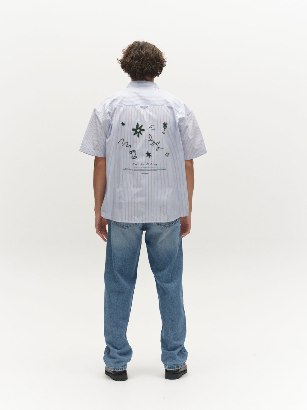A person faces away, wearing a light blue striped short-sleeve shirt with abstract black designs and text on the back, paired with Gabba GABmax Elvis Jeans in mid blue denim, set against a plain white background.