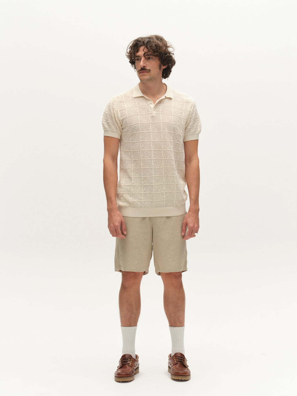 A man with wavy hair and a mustache stands against a plain white background, wearing the Gabba GABbenson Fiwi SS - Birch knit polo shirt, matching beige shorts, white socks, and brown boat shoes.