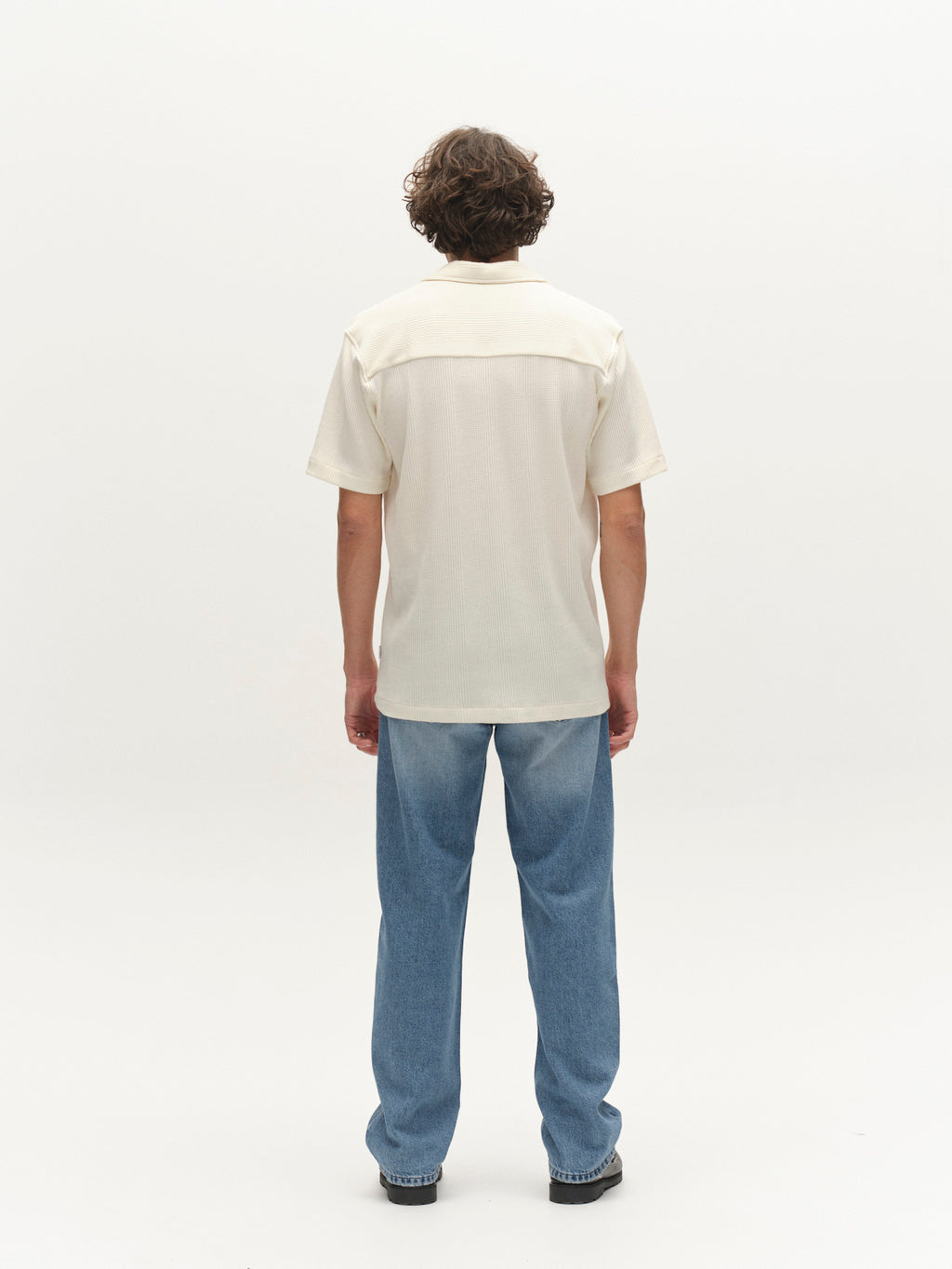 A person with curly hair faces away, wearing the Gabba GABvalente Wup SS T-Shirt in Bone White, light blue jeans, and black shoes against a plain white background.