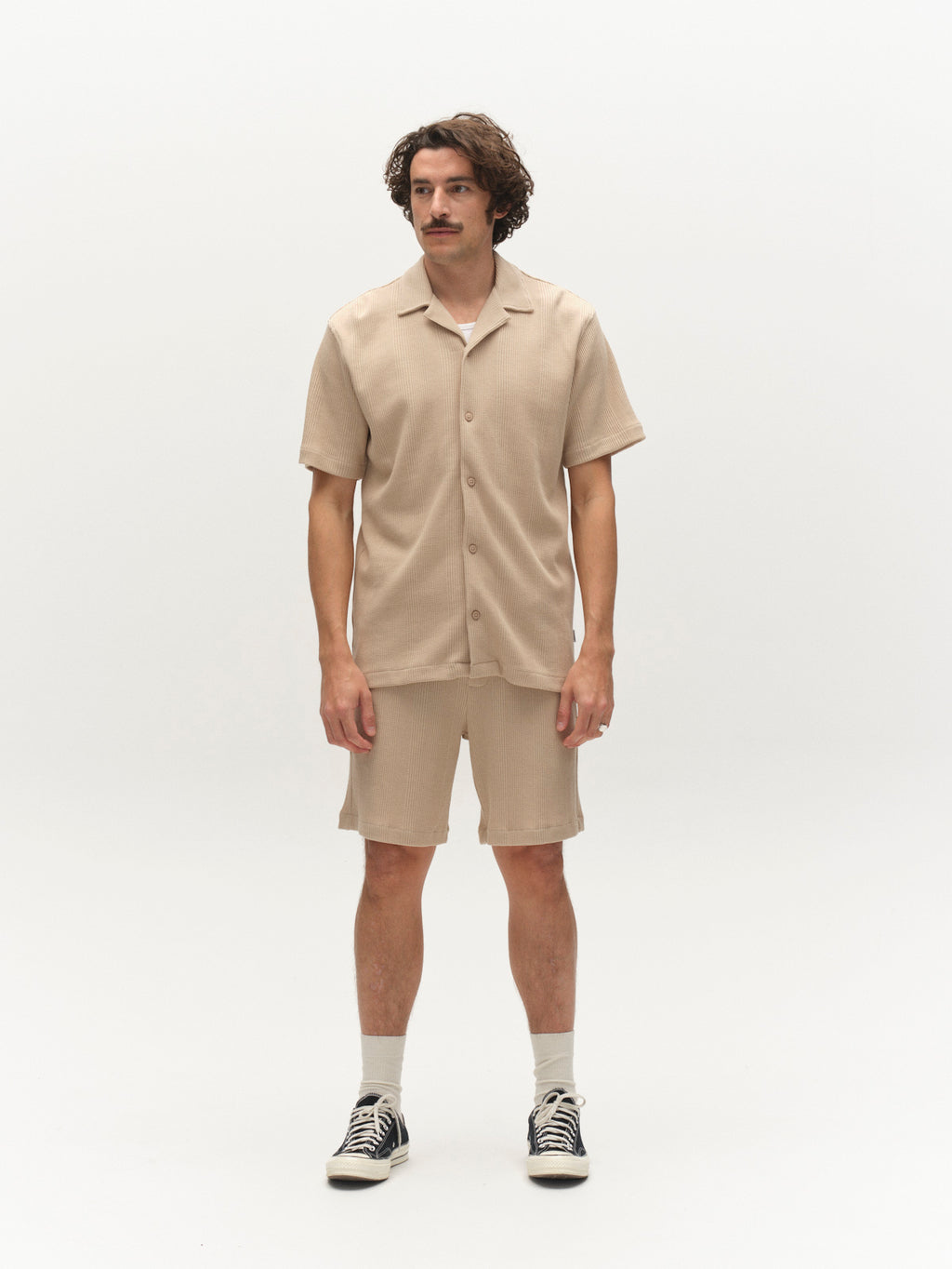 A man with wavy brown hair and a mustache stands against a white background, wearing the Gabba GABvalente Wup SS T-Shirt in Humus, matching beige shorts, white socks, and black sneakers.