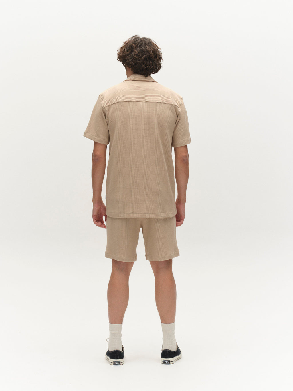 A person with short, wavy hair faces away from the camera, wearing a Gabba GABvalente Wup SS T-Shirt in Humus, matching beige shorts, white socks, and black shoes against a plain white background.