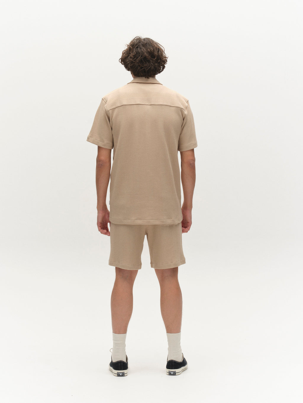 A person with short, wavy hair faces away, wearing the Gabba GABvalente Wup SS T-Shirt in Humus with matching shorts, white socks, and black sneakers against a plain white background.