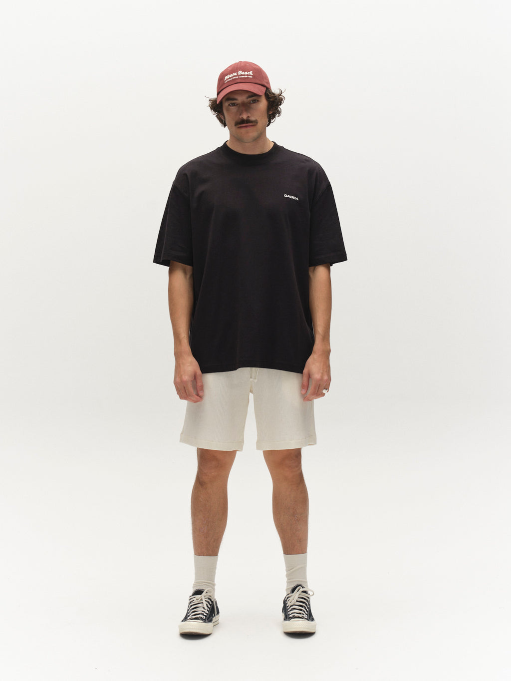 A person with a mustache and curly hair stands against a white background wearing GABtommi Wup Shorts in Bone White by Gabba, paired with a maroon cap, loose black T-shirt, white socks, and black-and-white sneakers.