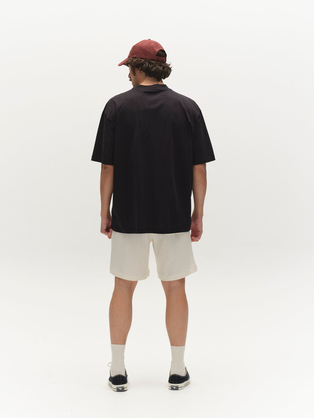 A man stands facing away, wearing a dark short-sleeve shirt, Gabba GABtommi Wup Shorts in Bone White, white socks, black slip-on shoes, and a red cap against a plain white background.