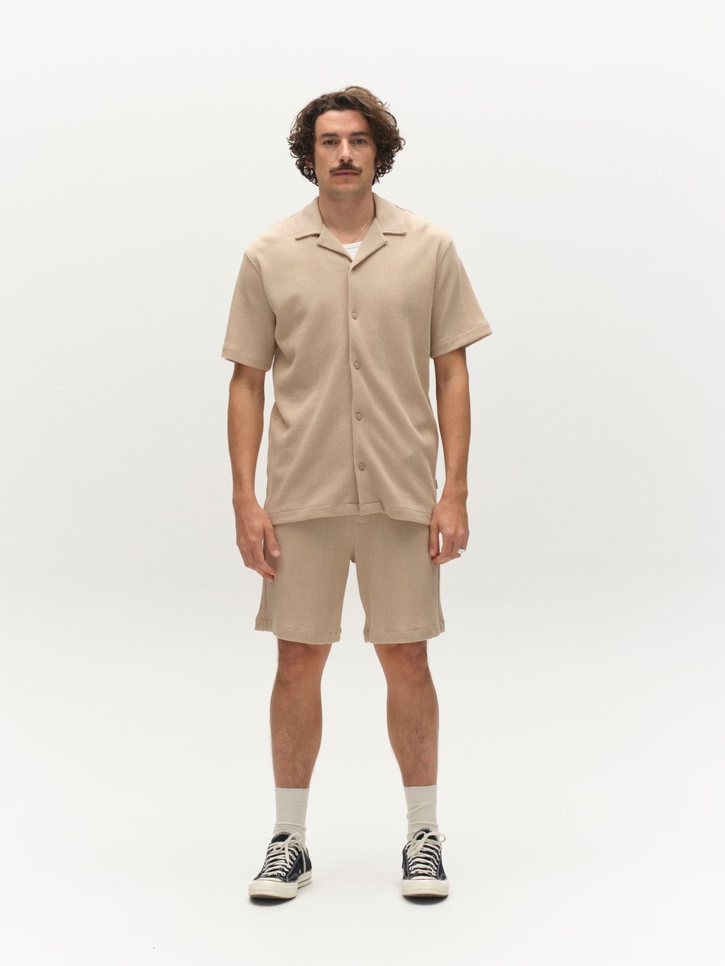 A man with wavy hair and a mustache stands facing forward against a plain white background, wearing a beige short-sleeve button-up shirt, Gabba GABtommi Wup Shorts - Humus, white socks, and black sneakers.
