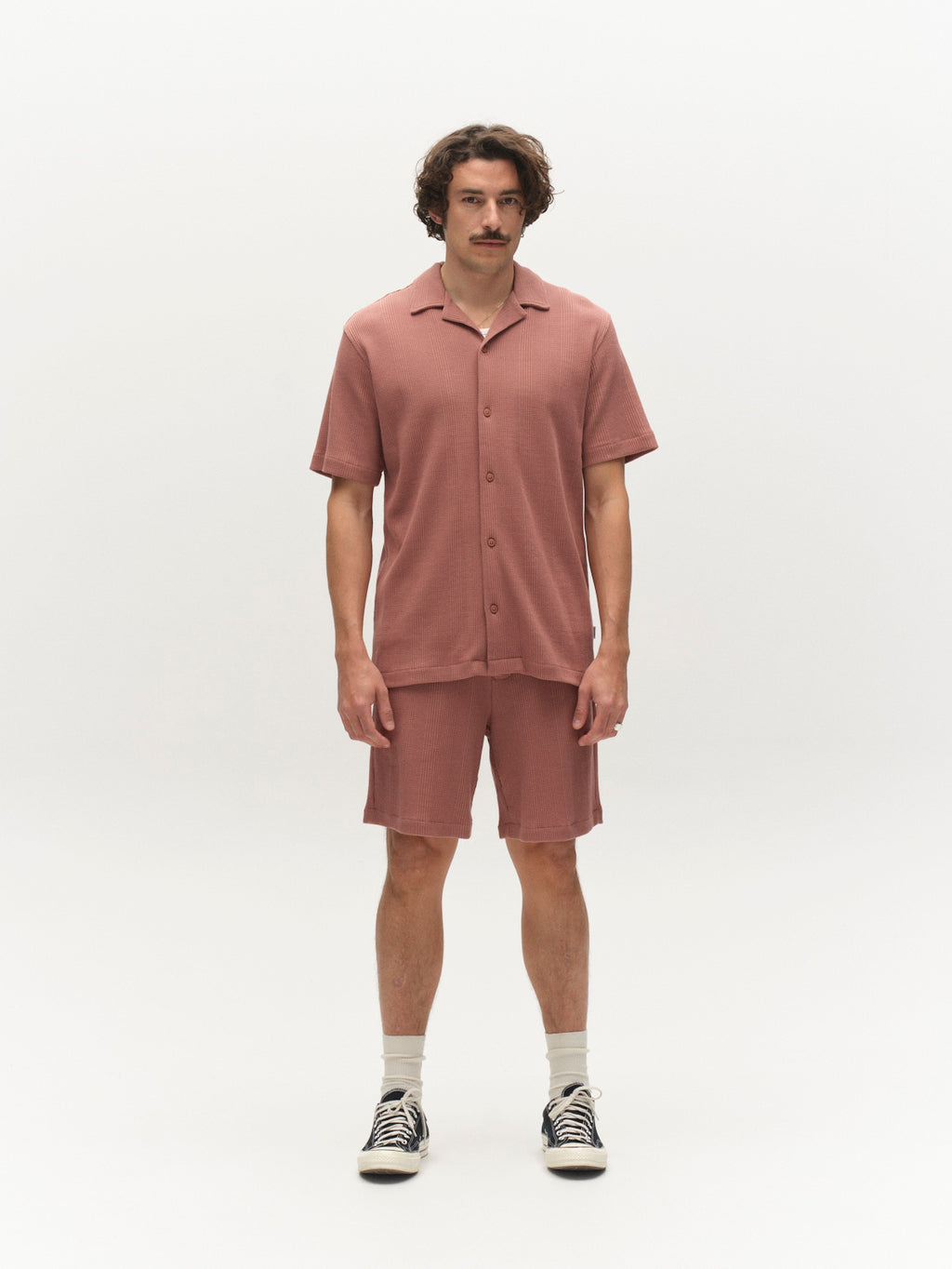 A man with wavy brown hair and a mustache stands facing forward against a white background, wearing Gabba GABtommi Wup Shorts in Wood Pink, a rose-colored shirt, white socks, and black high-top sneakers.