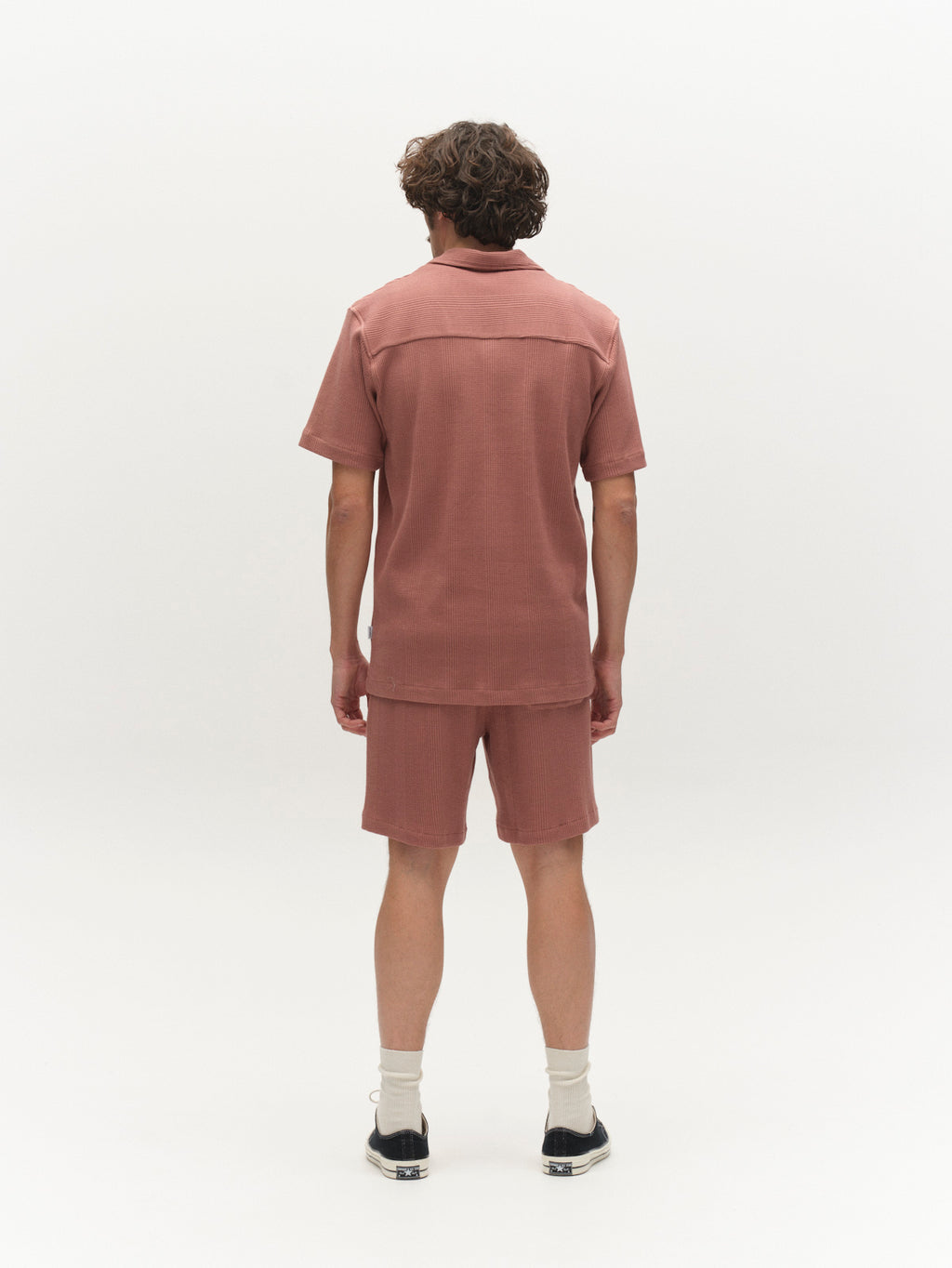 A person with curly hair is facing away, wearing Gabba’s GABtommi Wup Shorts in Wood Pink with a matching short-sleeve shirt, white socks, and black shoes against a plain white background.