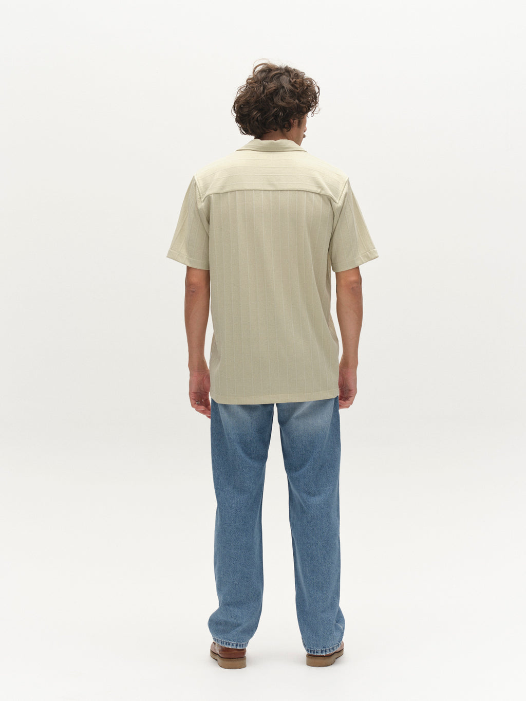 A person with curly hair faces away, wearing the Gabba GABvalente Armor SS T-Shirt in Alfa Green, paired with blue jeans and brown shoes, against a plain white background.