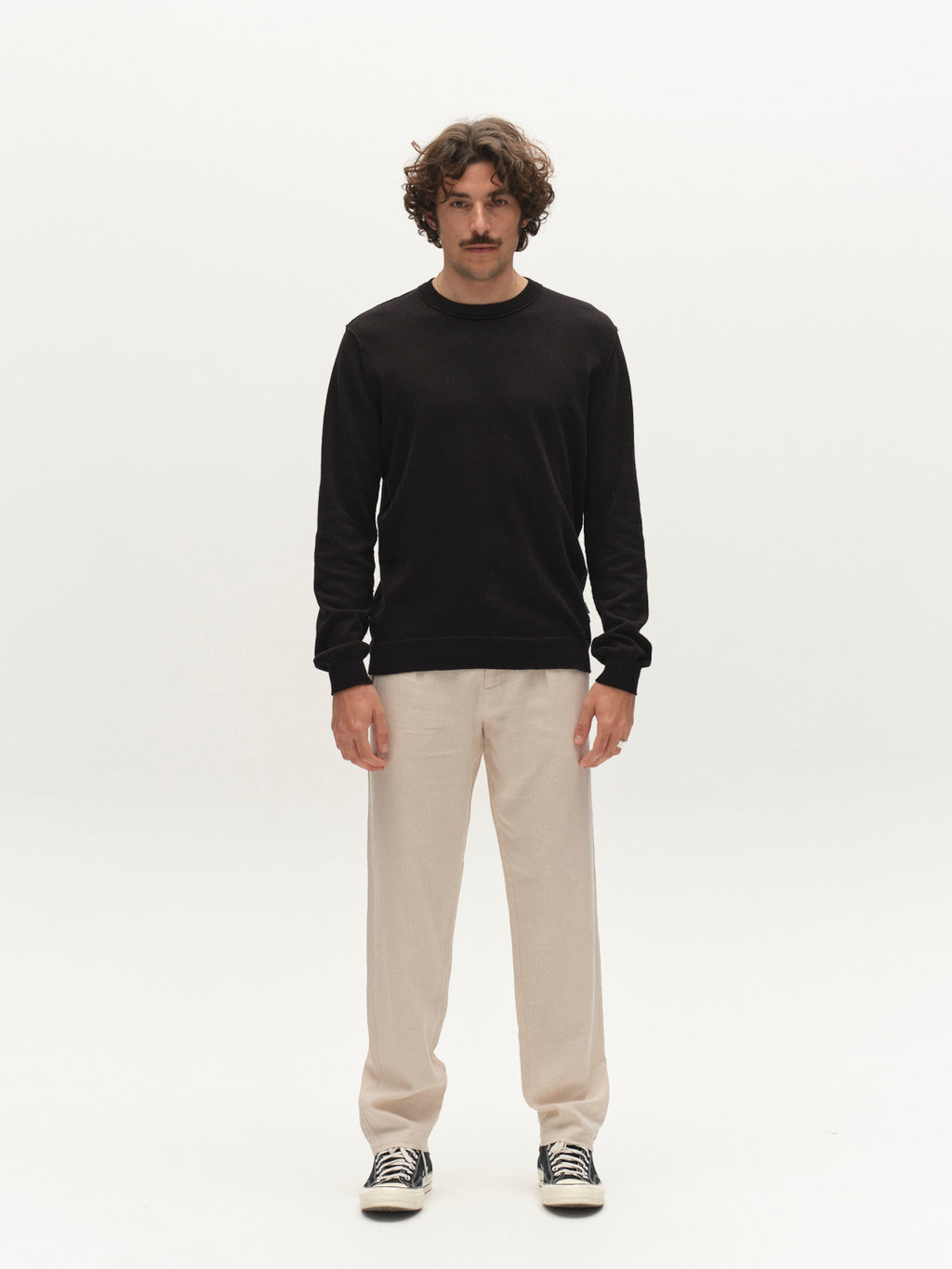 A man with curly hair and a mustache stands facing forward against a plain white background. He wears a black long-sleeve shirt, Gabba GABnapoli 19 Lino Pant in Birch, and black-and-white sneakers.