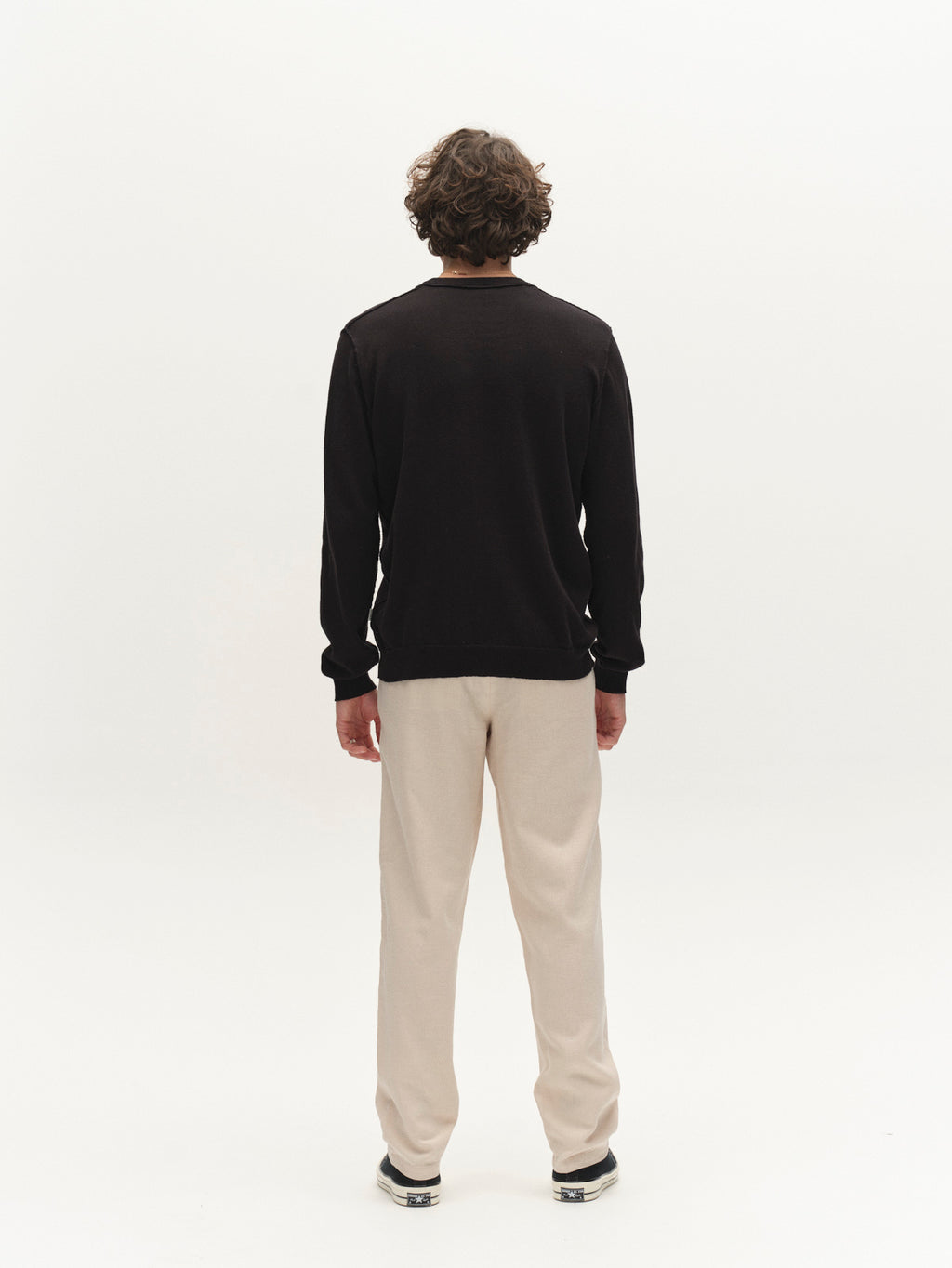 A person with curly hair stands facing away from the camera, wearing a black long-sleeve shirt, Gabba GABnapoli 19 Lino Pant in Birch, and sneakers, against a plain white background.