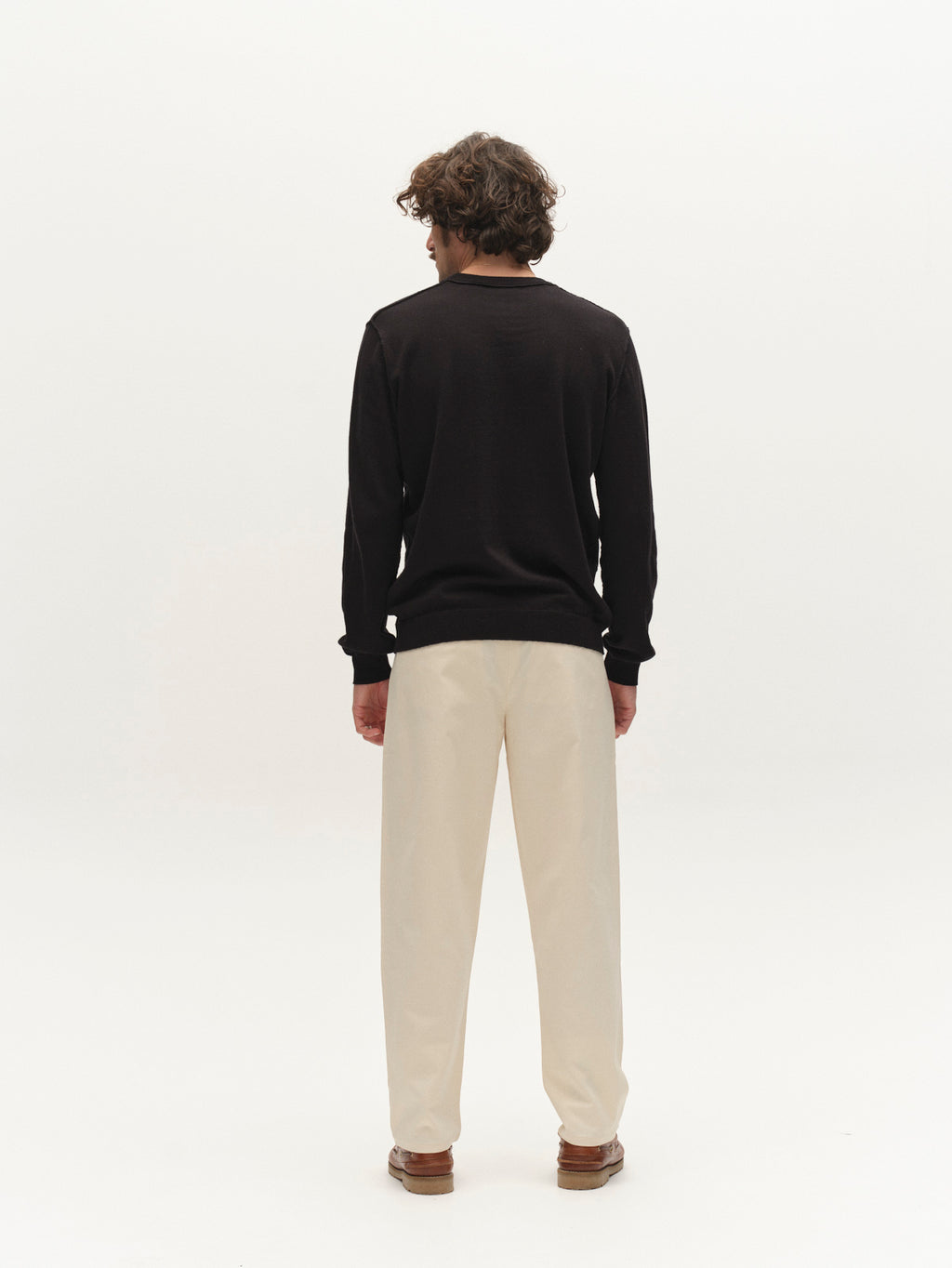 A person with curly hair wearing a black long-sleeve shirt, Gabba GABfirenze Bolu Cropped TENCEL™ Pant in Birch, and brown shoes stands facing away against a plain white background.