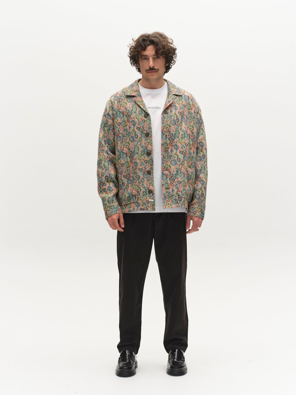 A man with curly hair and a mustache stands against a white background, wearing a colorful floral jacket over a white T-shirt, Gabba’s GABfirenze Bolu Cropped TENCEL™ Pant in black, and black shoes.