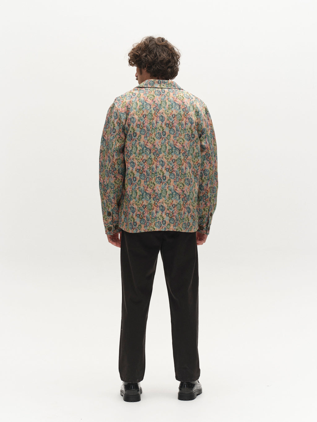 A person with curly hair, wearing a colorful floral-patterned jacket and the Gabba GABfirenze Bolu Cropped TENCEL™ Pant in black, featuring a relaxed fit, stands facing away on a plain white background.