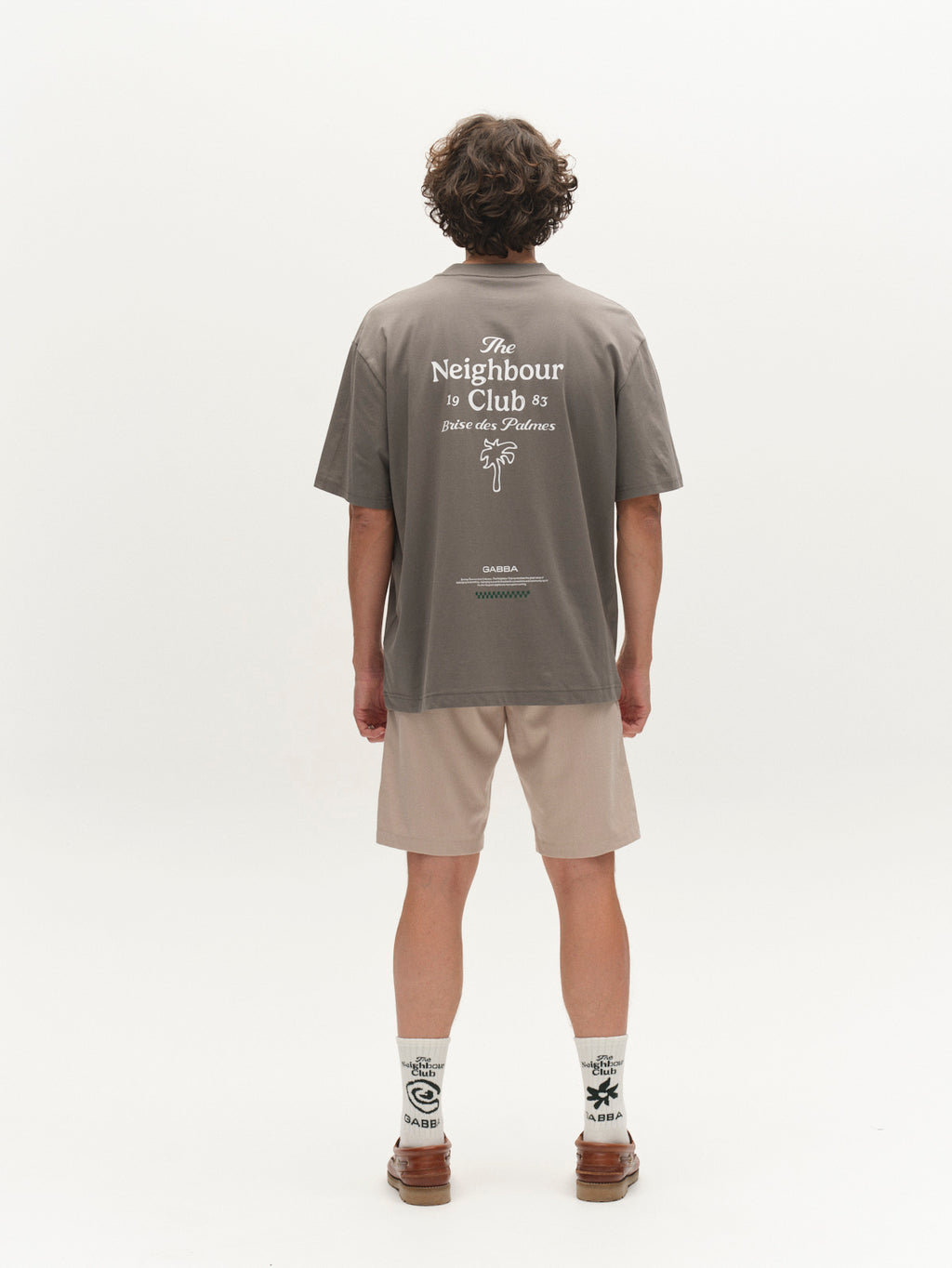 A person with curly hair stands facing away, wearing a gray T-shirt with white text, Gabba GABjet Deme Shorts in Birch, patterned socks, and brown shoes against a plain white background.