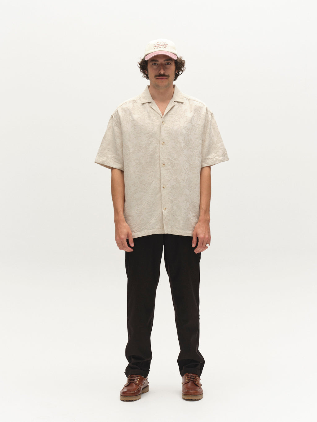 A man with a mustache stands against a white background, wearing a beige short-sleeve button-up shirt, Gabba GABjoe Turnup Fin Pant in black, brown shoes, and a light-colored baseball cap.