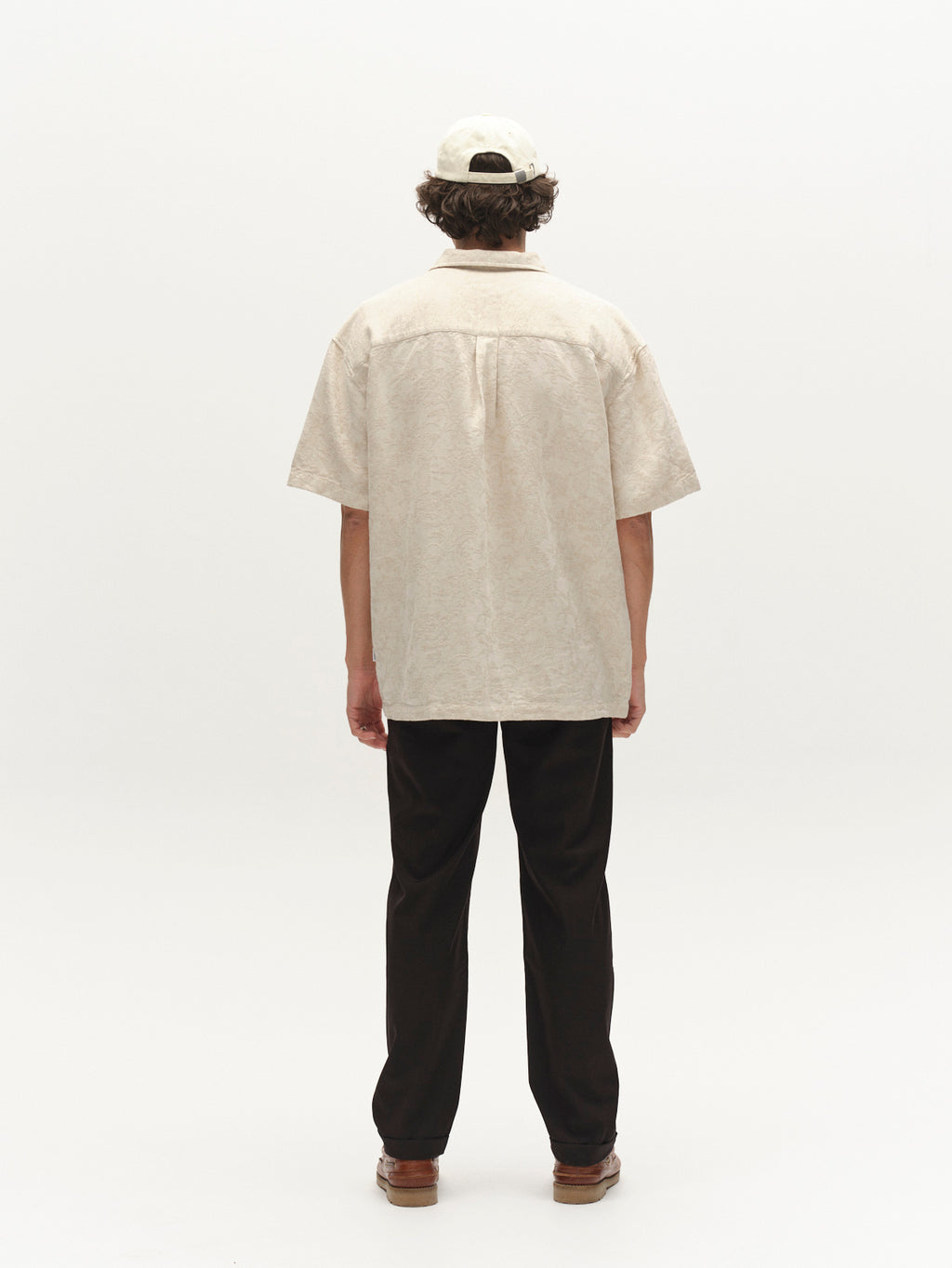 A person stands facing away from the camera in a cream short-sleeve shirt, brown shoes, a light baseball cap, and Gabba's GABjoe Turnup Fin Pant in black, set against a plain white background.