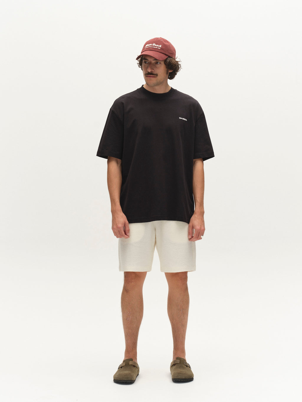 A person with a mustache stands against a plain white background, wearing a red cap, black t-shirt, brown slip-on shoes, and Gabba’s GABtommi Towel Shorts - Egret with an elasticated waistband.