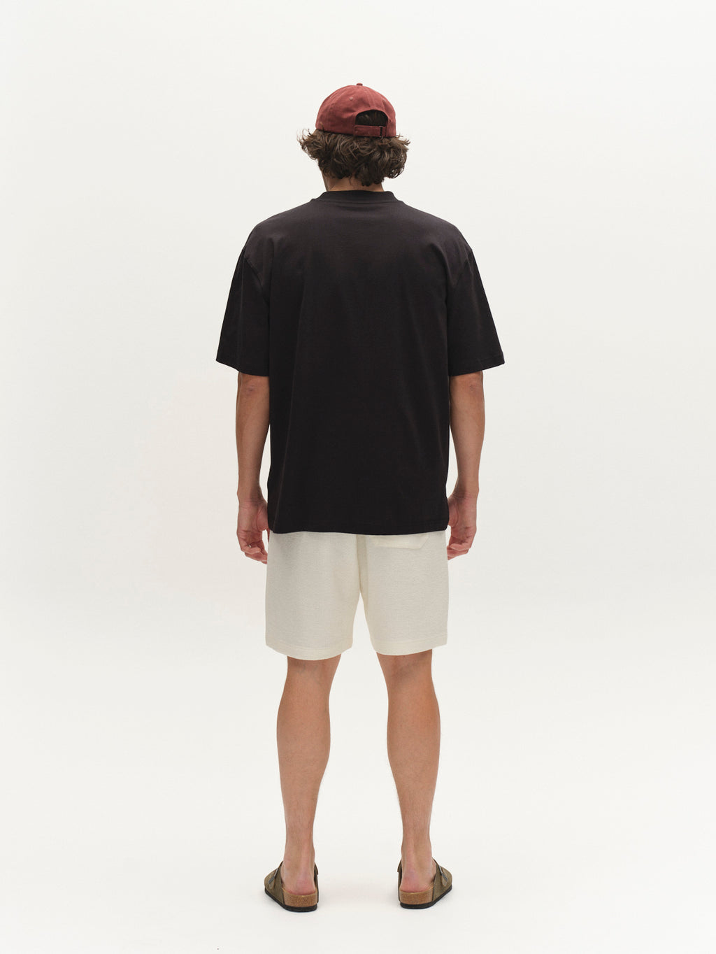 A person stands facing away from the camera, wearing a black T-shirt, Gabba GABtommi Towel Shorts - Egret with an elasticated waistband, sandals, and a red baseball cap. The background is plain white.