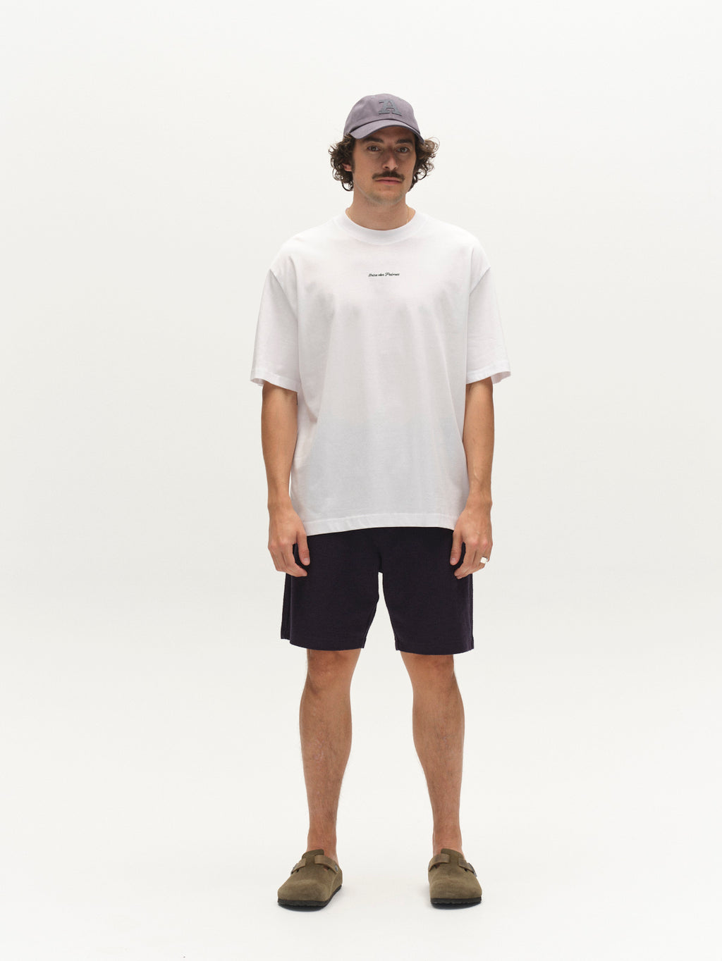 A man stands against a plain white background wearing a light purple baseball cap, loose white t-shirt, Gabba GABtommi Towel Shorts in navy with an elasticated waistband, and brown slip-on shoes. He has a mustache and relaxed hands at his sides.