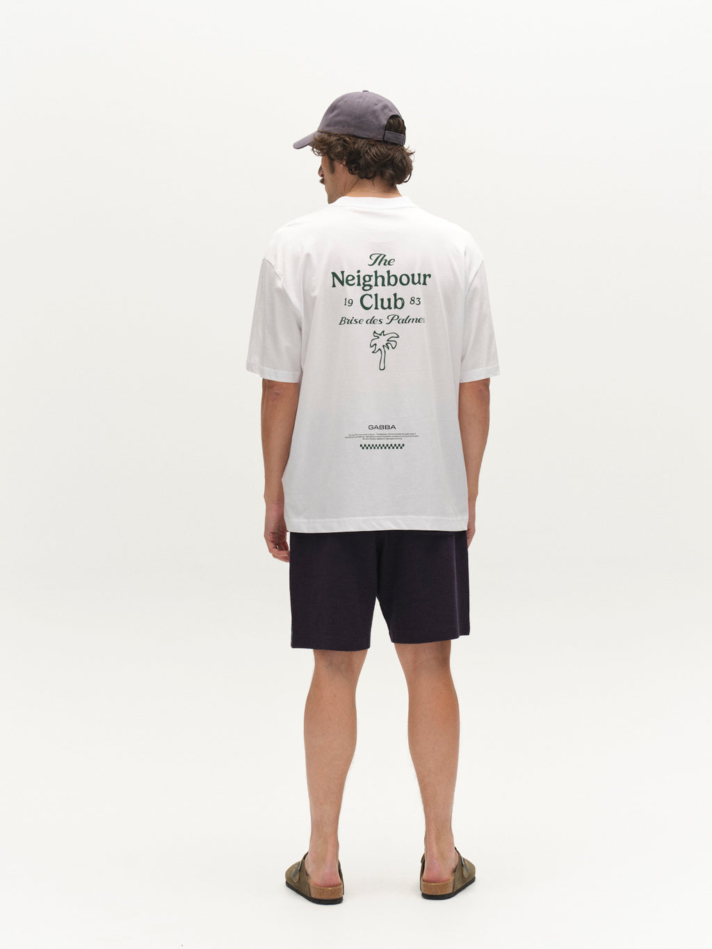 A person stands facing away, wearing a white T-shirt with green text and graphic, GABtommi Towel Shorts - Navy by Gabba featuring an elasticated waistband, a grey baseball cap, and sandals.