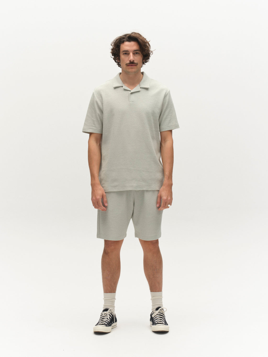 A man with wavy brown hair and a mustache stands against a white background, wearing a light gray polo shirt, Gabba’s GABtommi Towel Shorts in Mineral Gray, white socks, and black sneakers.