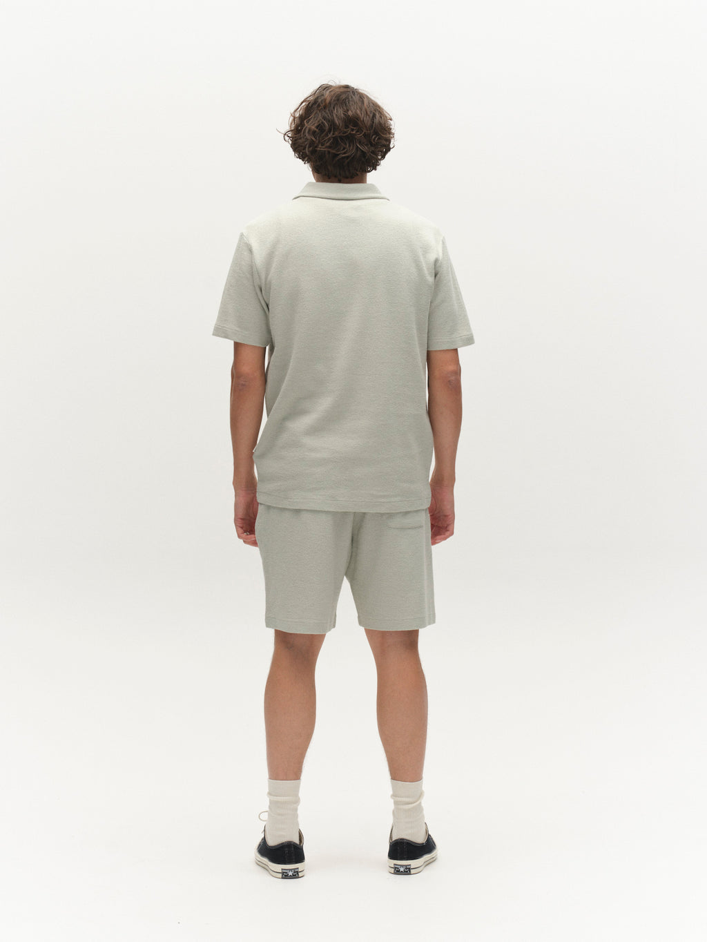 A person with short curly hair faces away from the camera, wearing a light gray polo, Gabba GABtommi Towel Shorts in Mineral Gray, white socks, and black sneakers against a plain white background.