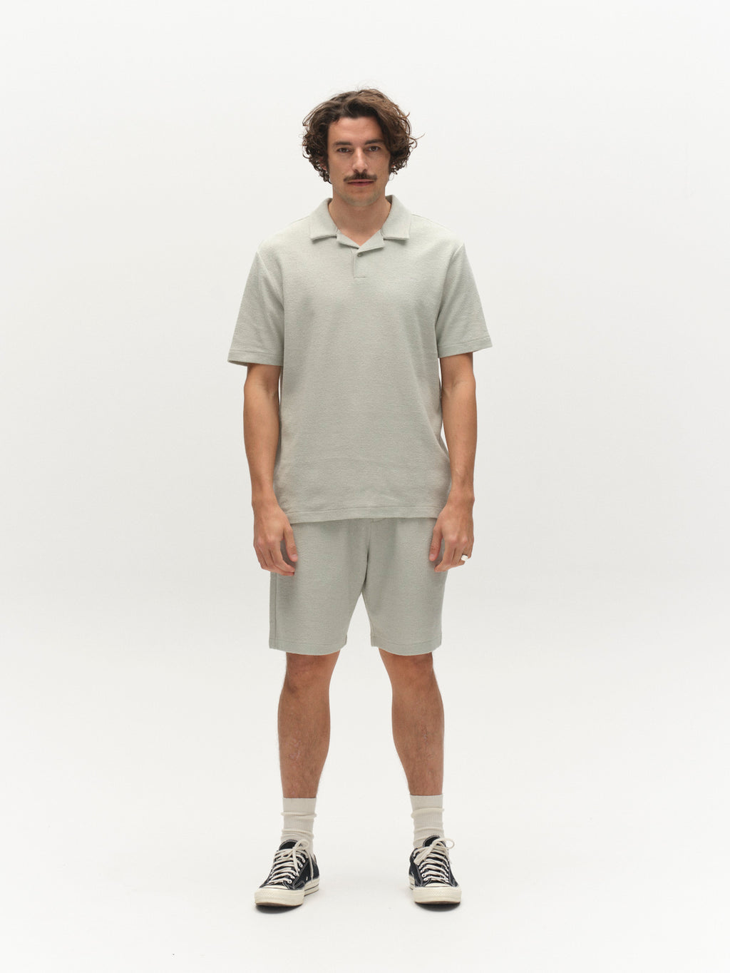 A man with wavy hair and a mustache stands facing forward in a Gabba GABlio Sku Towel T-Shirt - Mineral Gray, matching shorts, white socks, and black sneakers against a plain white background.