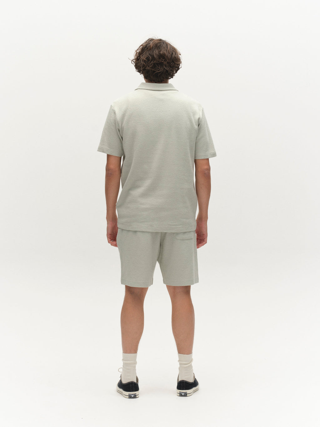 A person with short, curly hair is seen from behind wearing the Gabba GABlio Sku Towel T-Shirt in Mineral Gray with matching shorts, white socks, and black-and-white shoes against a plain white background.