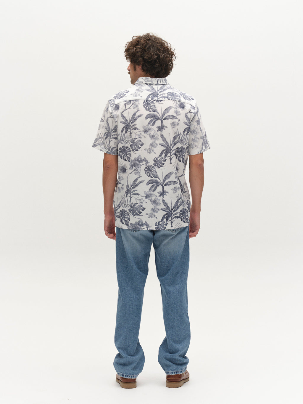 A person with curly hair stands facing away, wearing the Gabba GABsven Reg Leaves SS Shirt - Flower Mix and light blue jeans. The short-sleeve floral shirt is made from piqué jersey cotton. The background is plain white.