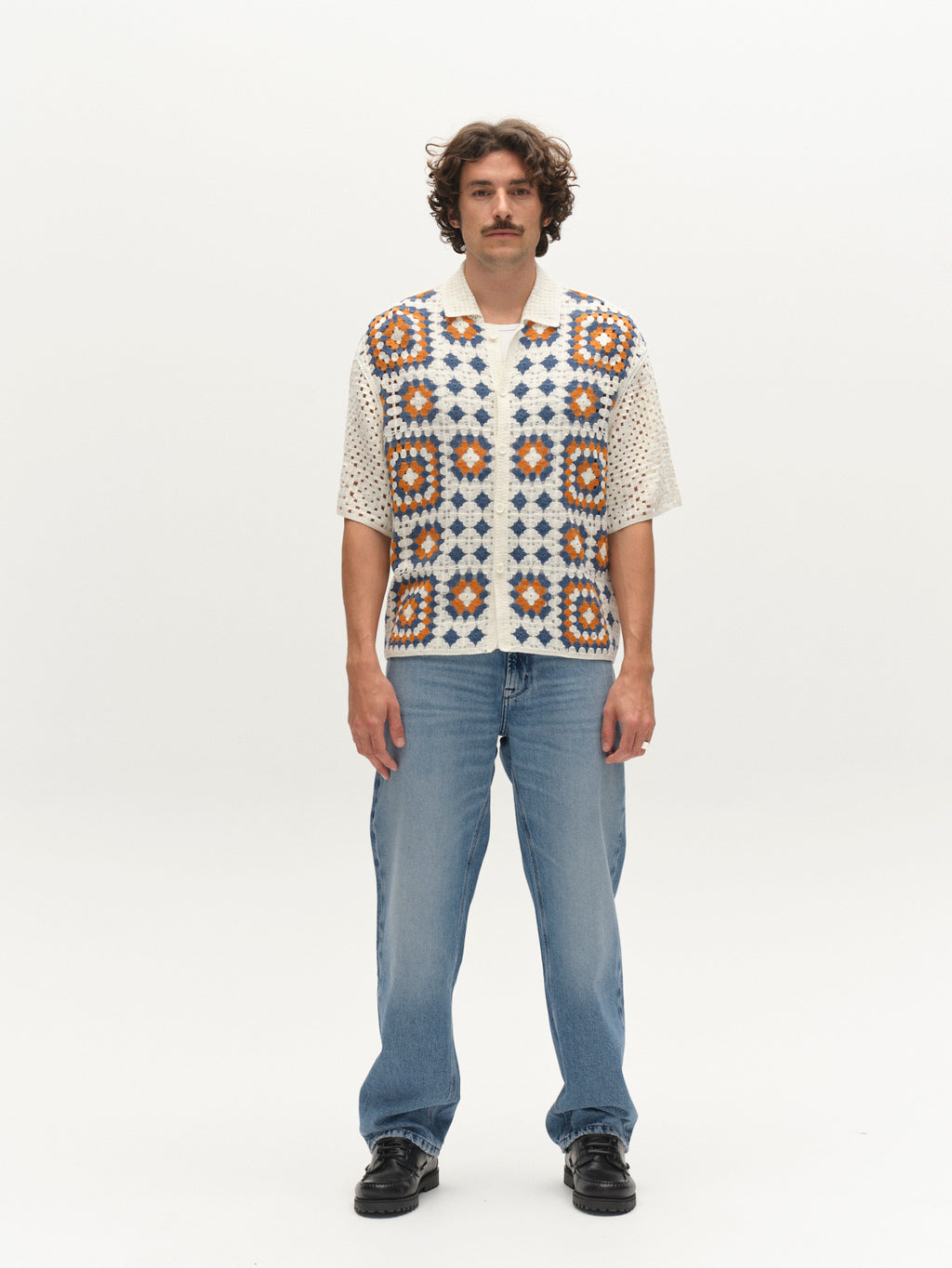 A man with curly hair and a mustache faces forward, wearing the Gabba GABcrochet Pattern SS Knit - Multi Pattern over a cream short-sleeve shirt, blue jeans, and black shoes, set against a plain white background.