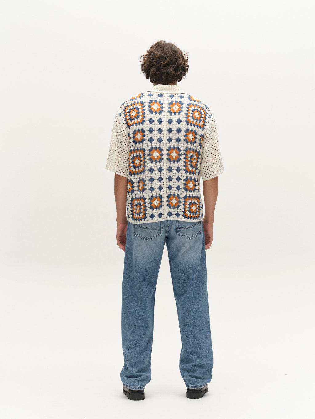 A person with curly hair faces away, wearing light blue jeans and the Gabba GABcrochet Pattern SS Knit - Multi Pattern shirt featuring a colorful geometric back, styled as a modern layer against a plain white background.
