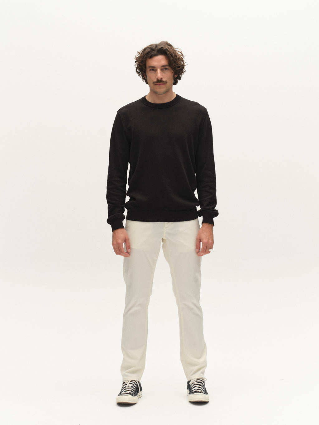 A person with curly hair and a mustache faces forward, wearing a black long-sleeve shirt, Gabba GABpaul K3280 Dale Chino - White pants, and black-and-white sneakers against a plain white background.