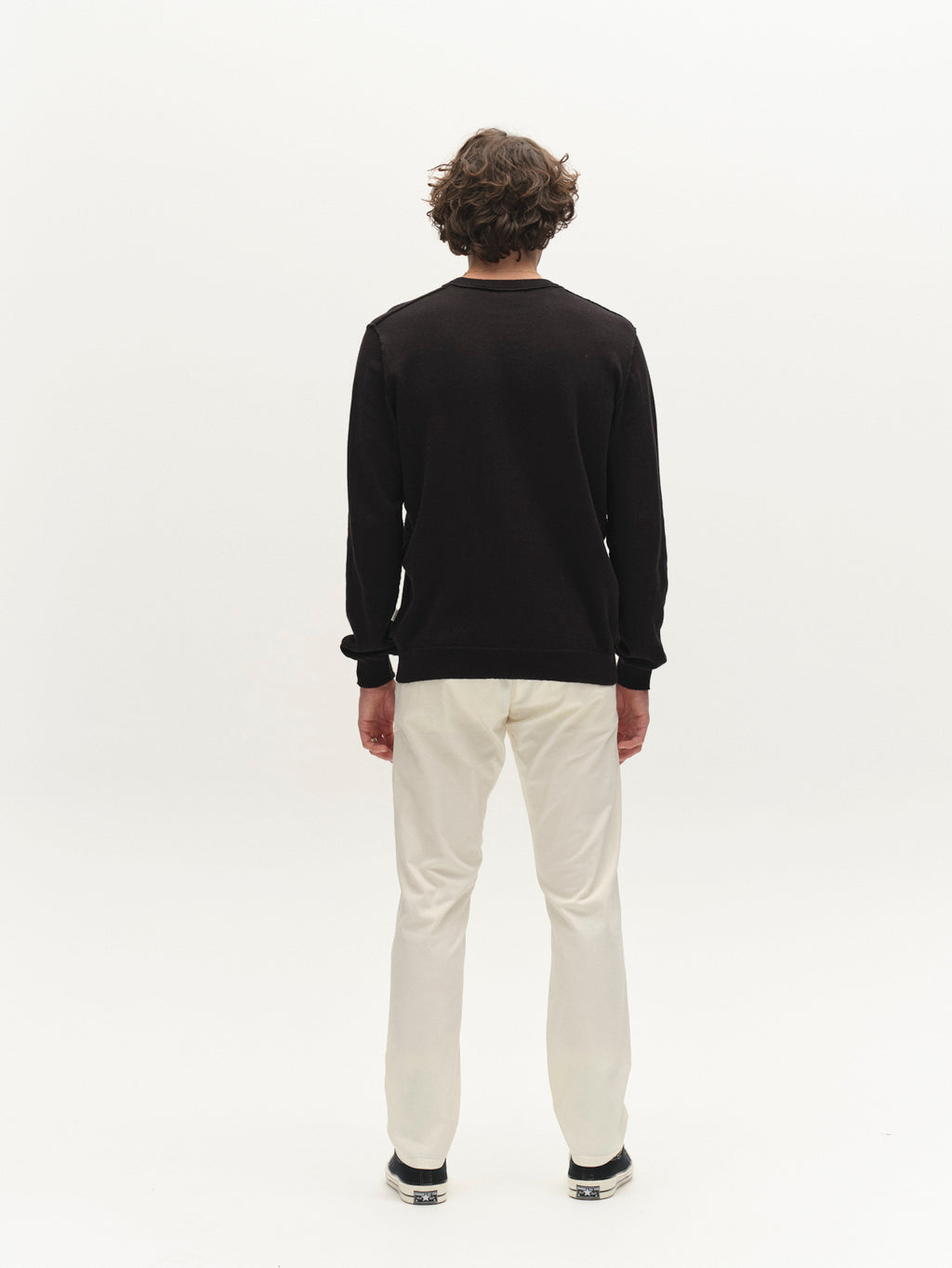 A person with wavy, brown hair stands facing away from the camera, wearing a black long-sleeve top and Gabba GABpaul K3280 Dale Chino - White pants with French pockets and black shoes against a plain white background.