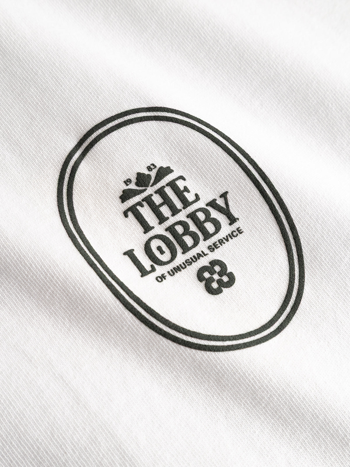 The Gabba GABlegin Boxy Fiore SS T-Shirt - White features a white textured cotton jersey fabric adorned with a black oval logo that reads “THE LOBBY OF UNUSUAL SERVICE” in capital letters, along with subtle decorative graphics above and below the text—an effortlessly cool contemporary wardrobe staple.