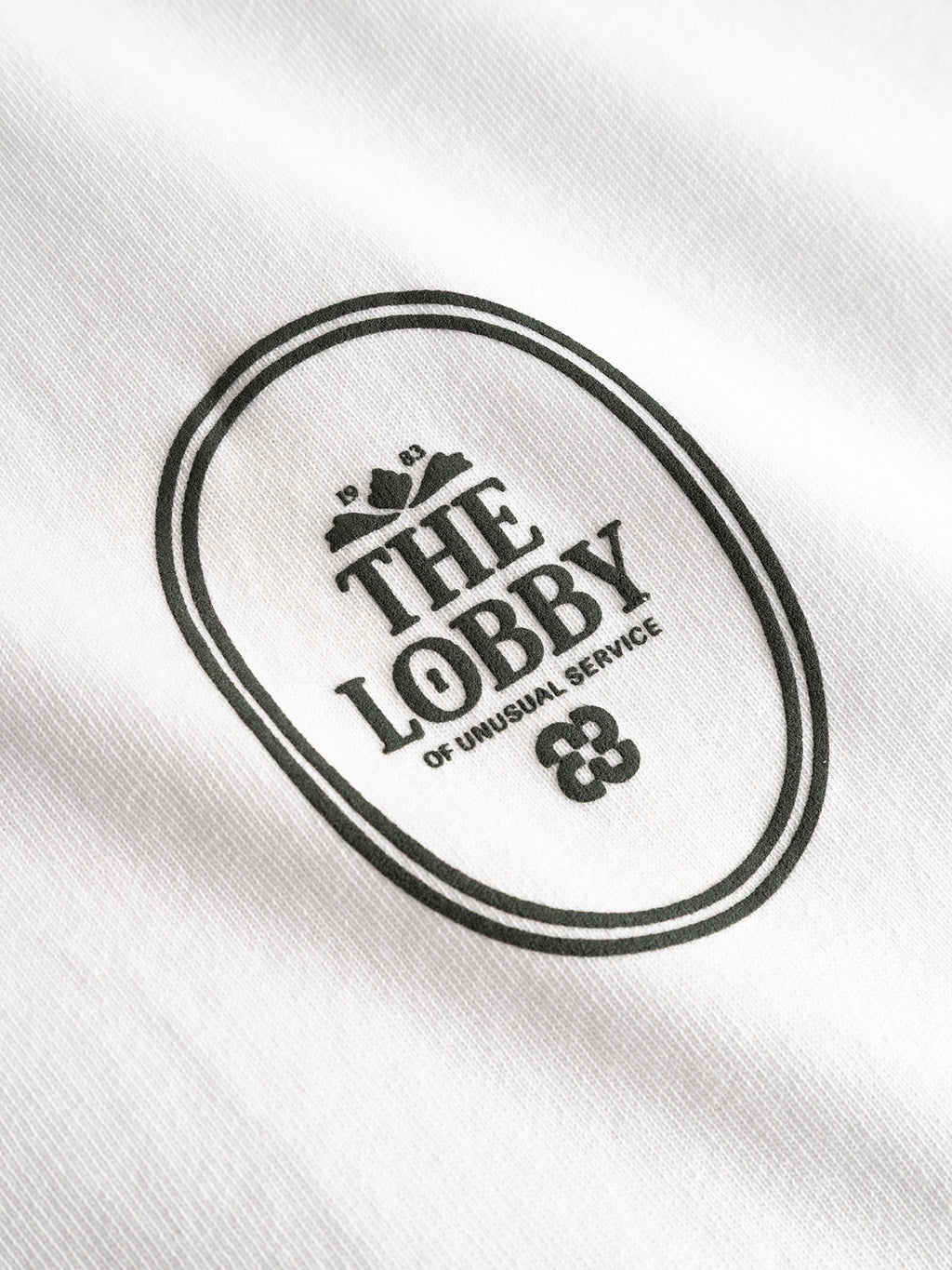 The Gabba GABlegin Boxy Fiore SS T-Shirt - White features a white textured cotton jersey fabric adorned with a black oval logo that reads “THE LOBBY OF UNUSUAL SERVICE” in capital letters, along with subtle decorative graphics above and below the text—an effortlessly cool contemporary wardrobe staple.
