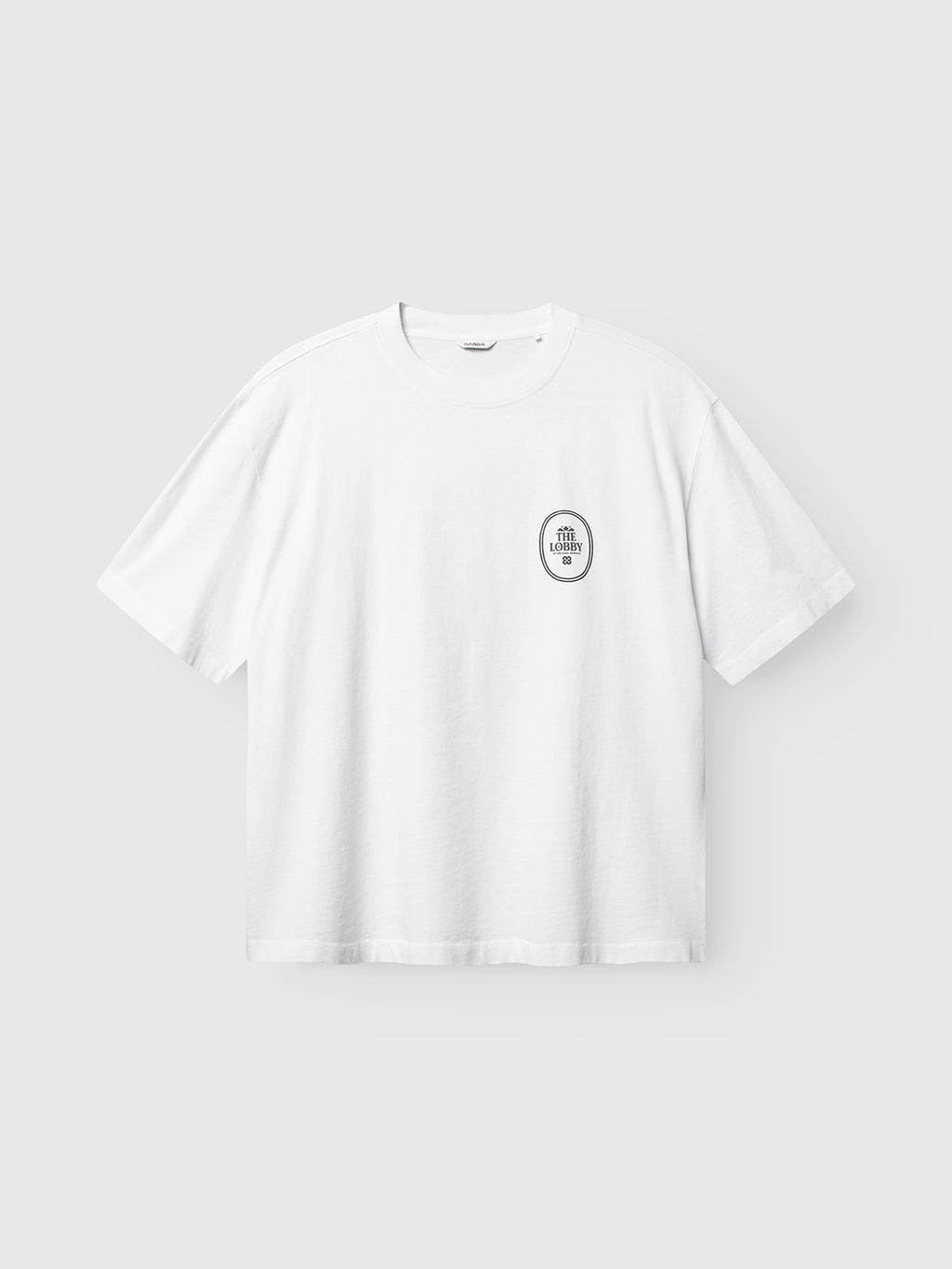 Introducing the Gabba GABlegin Boxy Fiore SS T-Shirt in white—a modern wardrobe essential. This short-sleeve t-shirt showcases a relaxed, boxy fit and is crafted from textured cotton jersey. A distinctive black and white round patch on the left chest reads “STAY CLOSE TO NATURE.” The shirt is presented against a light gray background.
