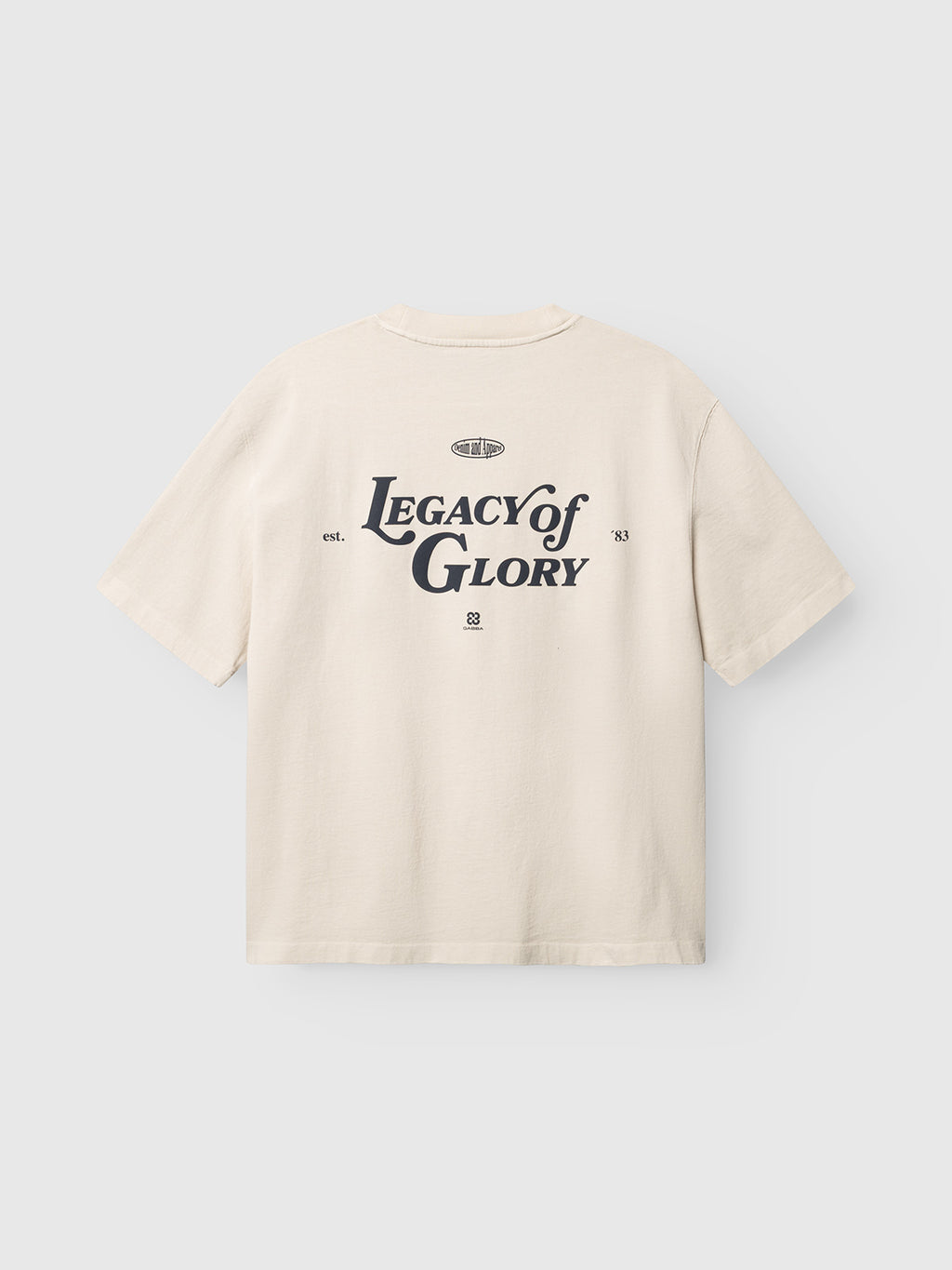The GABlegin Boxy Glory SS T-Shirt - Birch by Gabba is a beige short-sleeve t-shirt in a relaxed, boxy fit, crafted from textured cotton jersey. It features an eye-catching “Legacy of Glory” graphic printed in large black letters on the back, complemented by smaller decorative text and symbols.