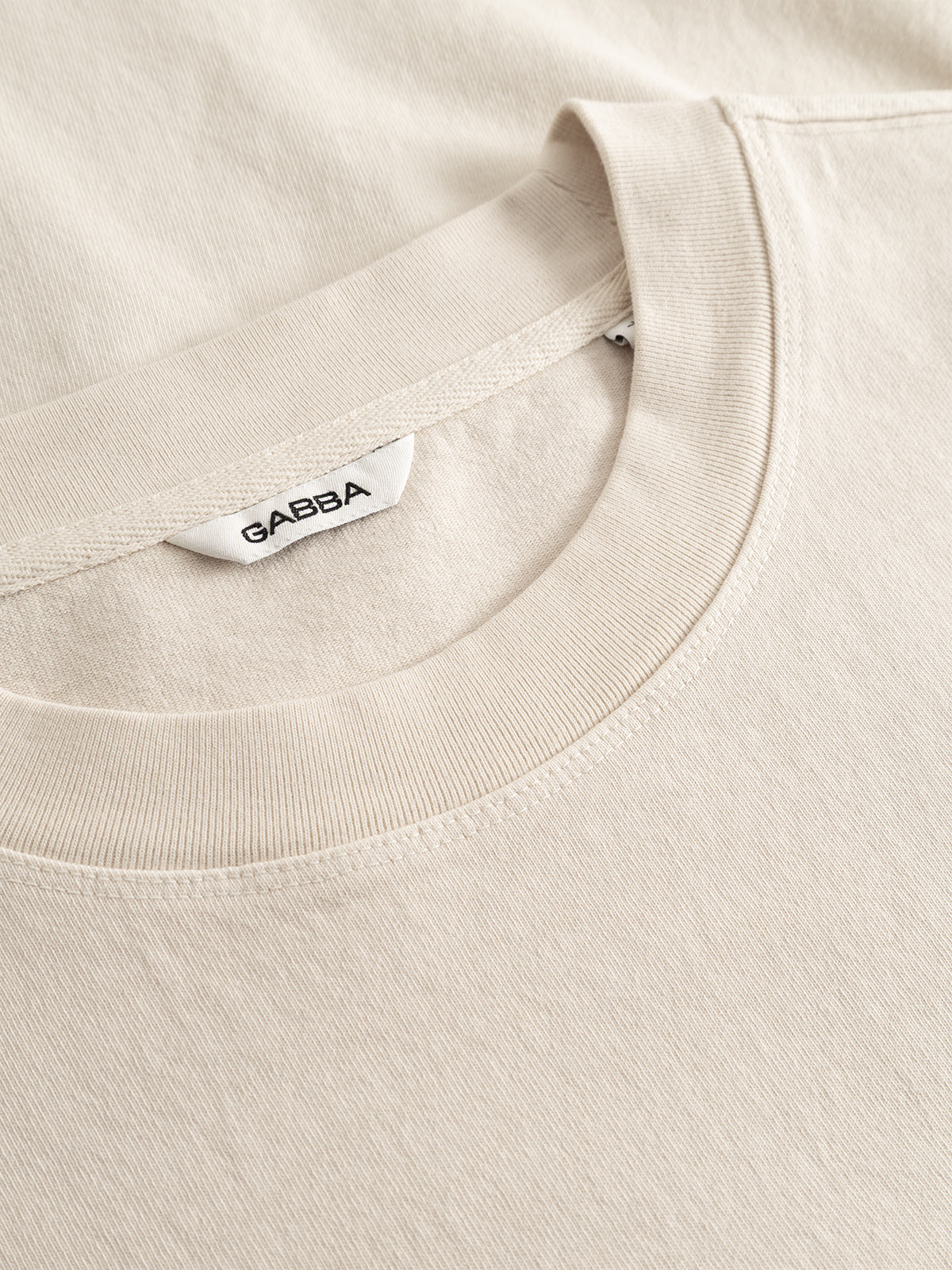 Close-up of the GABlegin Boxy Glory SS T-Shirt in Birch by Gabba, featuring a beige crew neck design with a white Gabba tag, crafted from textured cotton jersey and neatly laid flat on a matching beige surface.