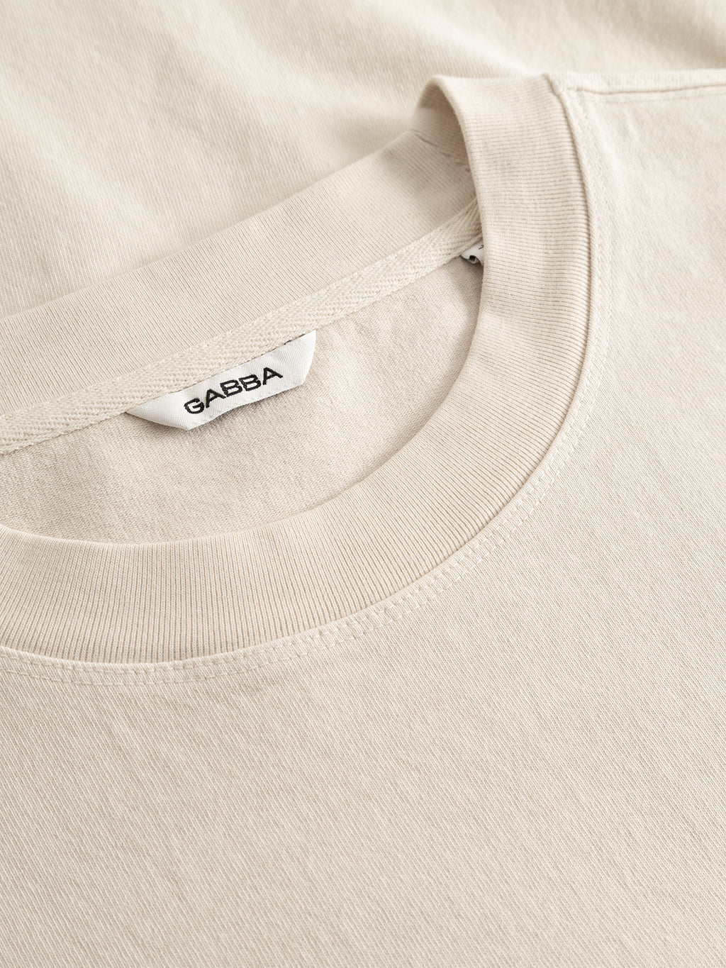 Close-up of the GABlegin Boxy Glory SS T-Shirt in Birch by Gabba, featuring a beige crew neck design with a white Gabba tag, crafted from textured cotton jersey and neatly laid flat on a matching beige surface.