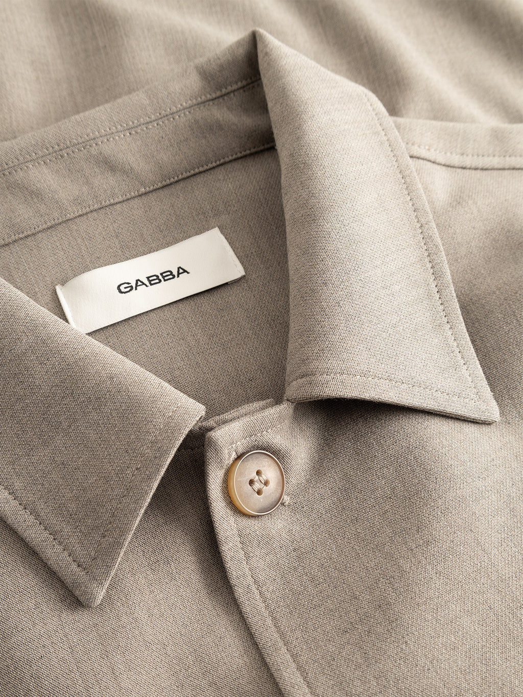 A close-up of the GABtogger Push Phin LS Overshirt GRS in Sand, featuring a beige collared design with a button and a white label displaying the Gabba brand name. Crafted from a wool-viscose blend, this regular fit overshirt is an essential layering piece for any modern wardrobe.