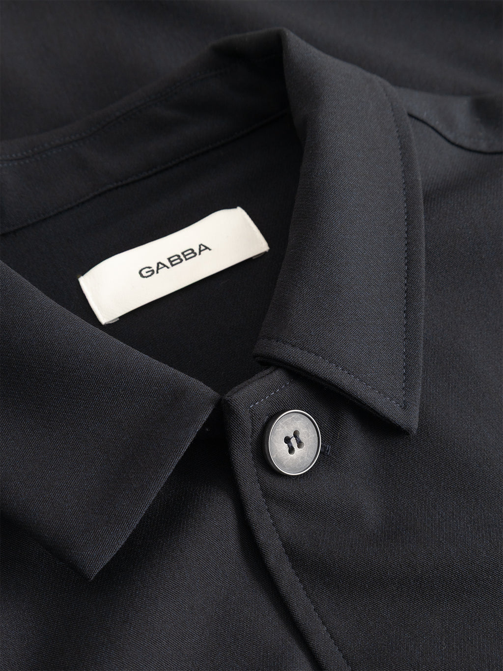 Close-up of the GABtogger Push Phin LS Overshirt GRS in Blue Black by Gabba—a dark-hued, collared shirt expertly crafted from a premium wool-viscose blend. A white label marked “GABBA” and a single grey button near the collar are visible, making this piece an ideal layering essential for any wardrobe.