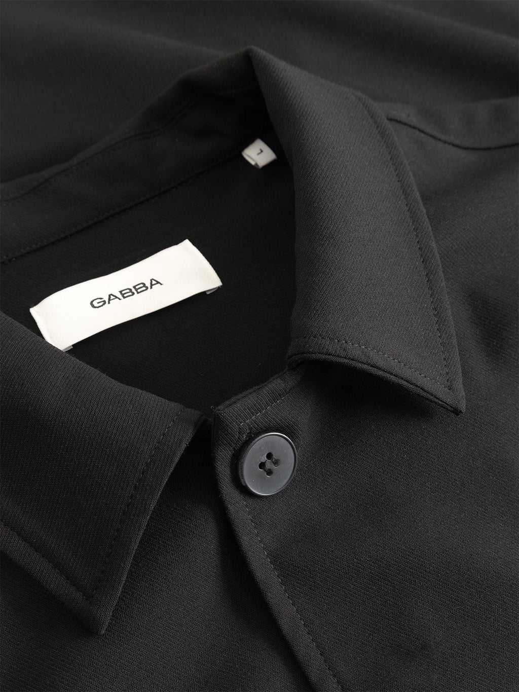 Close-up of the GABtogger Push Phin LS Overshirt GRS in black by Gabba, featuring a black collar with a button and an inner white tag labeled Gabba. Crafted from a soft wool-viscose blend, this overshirt is neatly laid out on a flat surface.