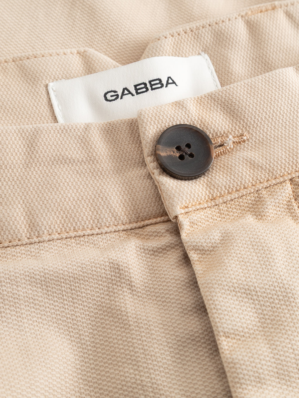 Close-up of the GABfirenze Cropped Marlo Pant in Birch by Gabba, featuring beige relaxed-fit trousers made from garment-dyed dobby fabric, a dark button, and a white label with the Gabba logo inside the waistband.