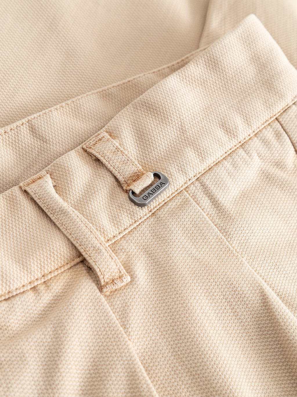 Close-up of the GABfirenze Cropped Marlo Pant in Birch by Gabba, showcasing the beige garment-dyed dobby fabric with a textured finish. The image highlights a belt loop and a small silver tag with engraved lettering, focusing on the waistband and upper front section of these relaxed fit trousers.