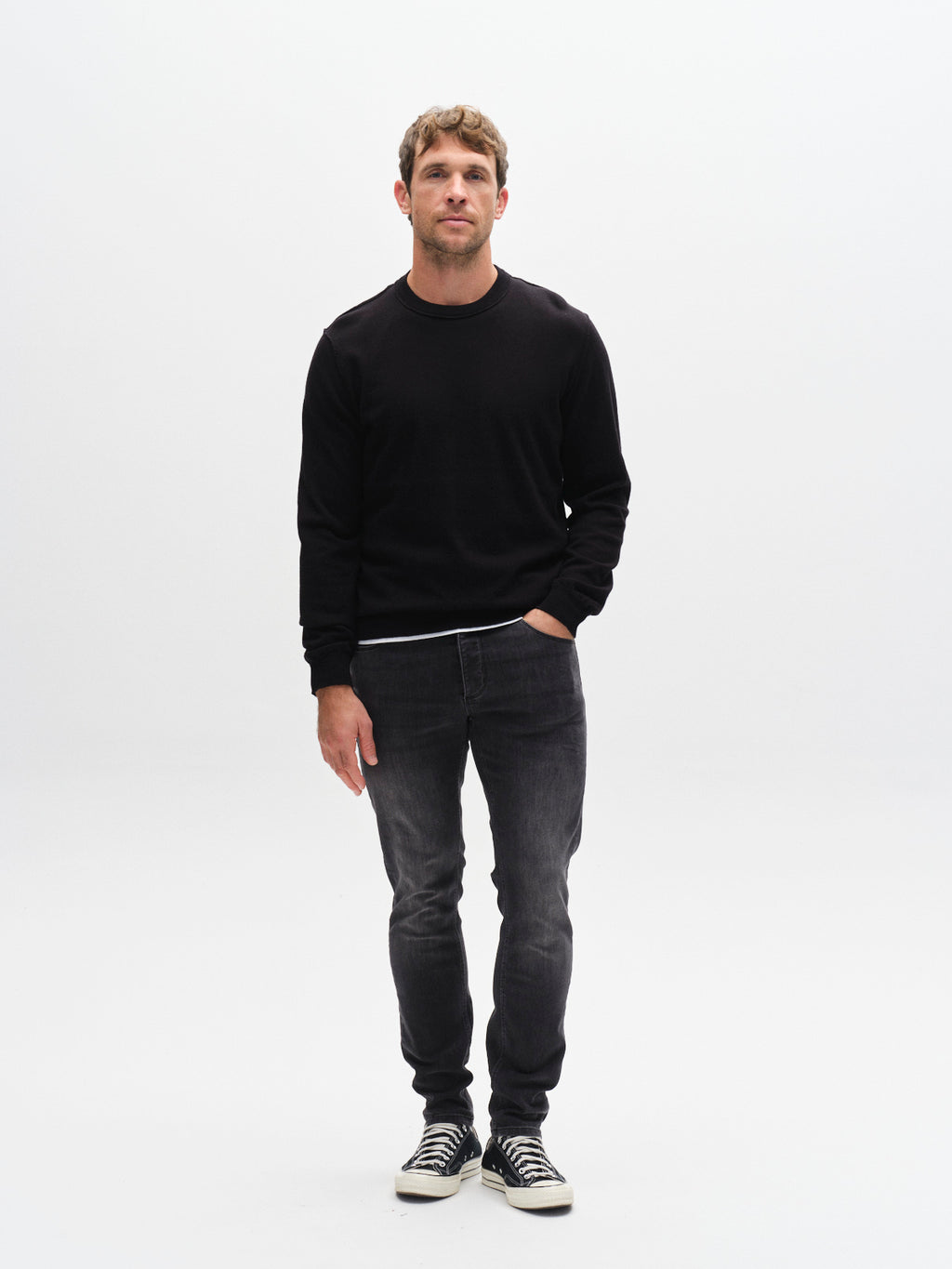 A man stands against a white background wearing a black sweater, GABBA GABrey Thor Jeans in Denim wash, and black-and-white sneakers. One hand is in his pocket as he looks at the camera with a laid-back vibe.