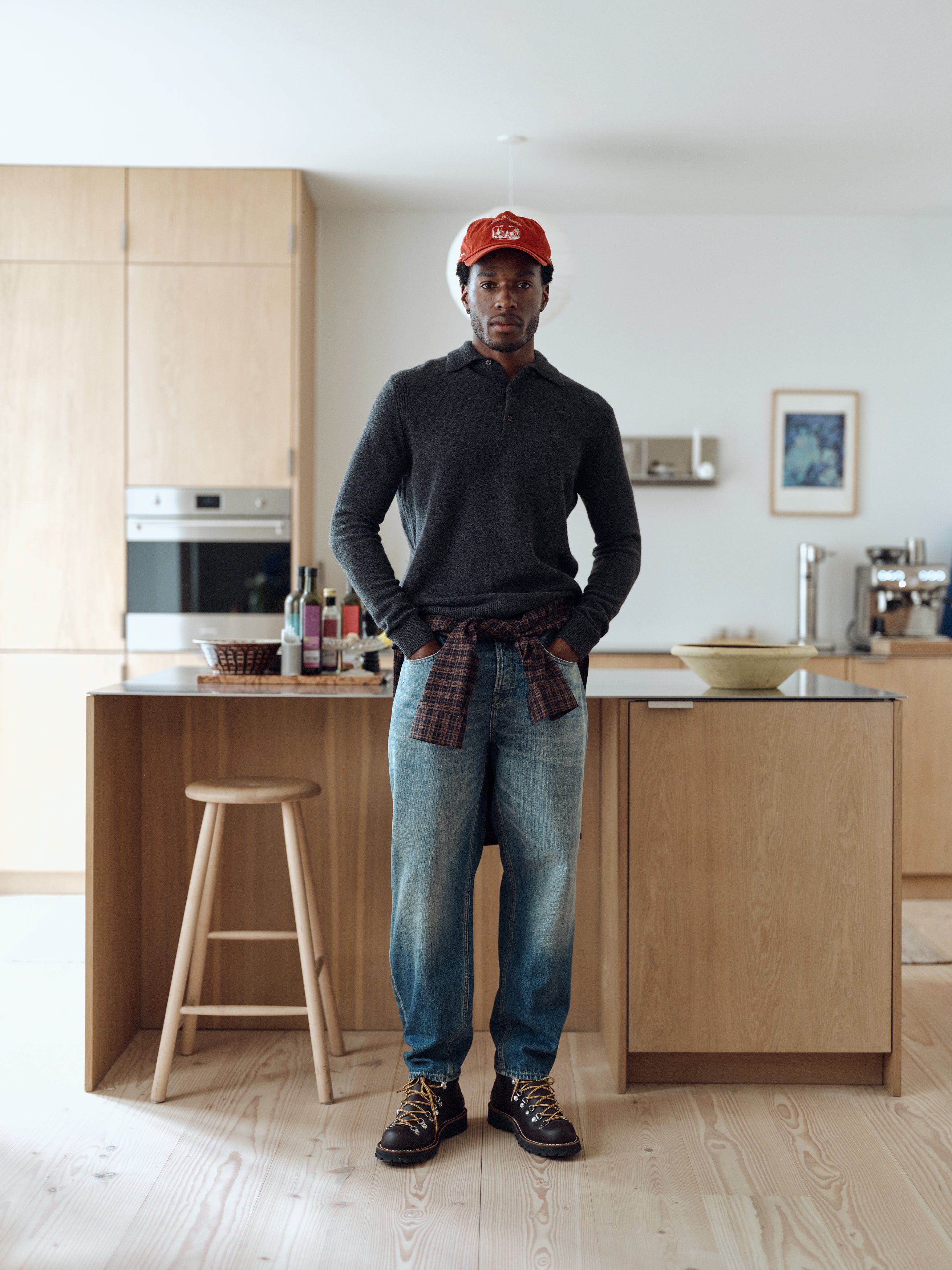 A man in a red cap, jeans, and boots stands in a modern kitchen with light wood cabinets and a stool. He is wearing the Gabba GABworren Polo Aico LS Knit in Dark Grey Mel. Bottles sit on the countertop, and a framed photo hangs on the white wall.