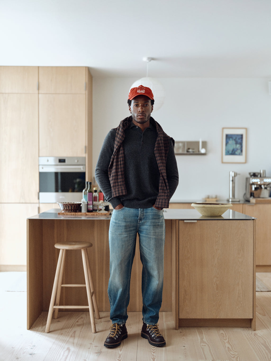 A man stands in a modern kitchen with wooden cabinetry, wearing jeans and a dark sweater. Draped over his shoulders is the Gabba GABsven Reg TX2 LS Shirt in Brown Slate, which features a classic checked texture and is crafted from soft yarn-dyed cotton. Completing his outfit, he sports a red cap and keeps his hands in his pockets as he looks at the camera.